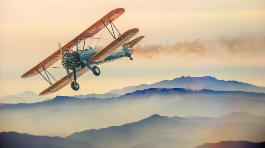 A vintage biplane in flight over misty mountains at sunset.